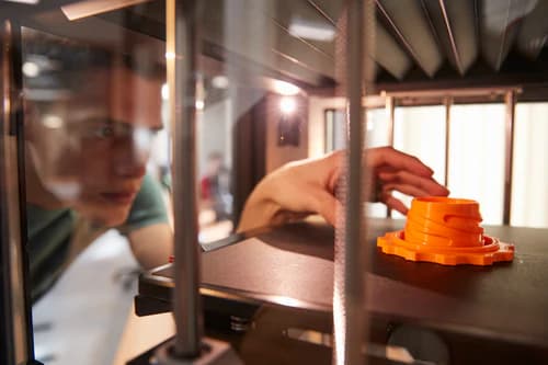 man reaching in to a 3D printer to remove an orange printed part